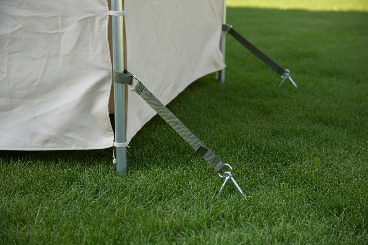 Abri de jardin sans permis : le pied d'un abri léger à toile, une sangle tendue reliant l'armature à un piquet d'ancrage planté dans la pelouse.