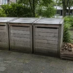 Trois bacs de compostage partagé au pied d'un immeuble, avec une réserve de matière sèche à côté