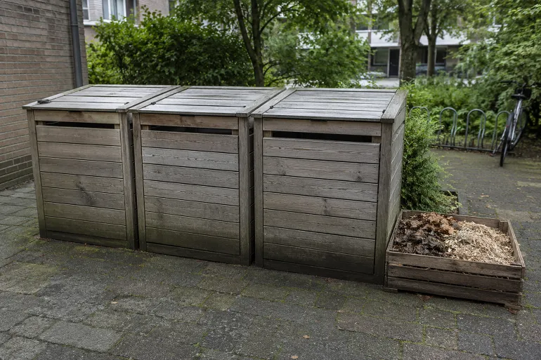 Trois bacs de compostage partagé au pied d'un immeuble, avec une réserve de matière sèche à côté