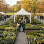 Fêtes et foires aux plantes 2026 : toutes les dates 5 Vue large d'une fête des plantes, un stand de pépiniériste professionnel avec des plantes alignées par variété dans des pots noirs identiques sous un barnum blanc, le producteur discutant avec des visiteurs.