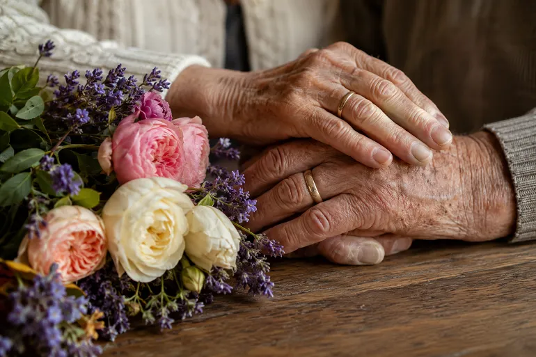 Les mains d'un couple âgé portant leurs alliances, posées côte à côte sur une table près d'un bouquet de roses et de lavande.