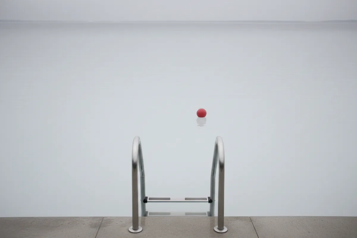 Eau trouble de piscine : vue rasante au ras de l'eau d'une piscine à l'eau opaque blanc-gris, une échelle inox entrant dans l'eau et disparaissant complètement après sa première marche immergée, sans aucun fond visible.