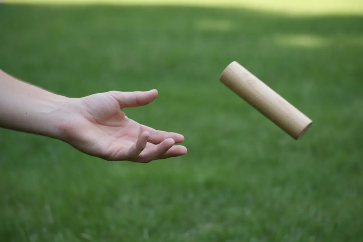 Un joueur lance un bâton de bois rond par en dessous, le bâton saisi en pleine rotation verticale bout sur bout au-dessus de la pelouse.