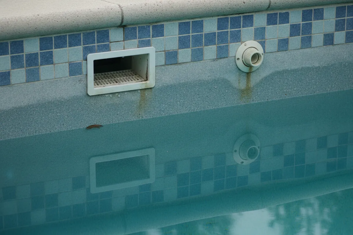 Gros plan sur une paroi de piscine où le niveau d'eau a été abaissé, laissant le skimmer et la buse de refoulement entièrement hors d'eau, avec une trace visible de l'ancien niveau sur le carrelage.