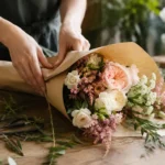 Deux mains emballent un bouquet de fleurs de saison dans du papier kraft sur un comptoir de bois, à côté de tiges coupées et d'une paire de ciseaux.