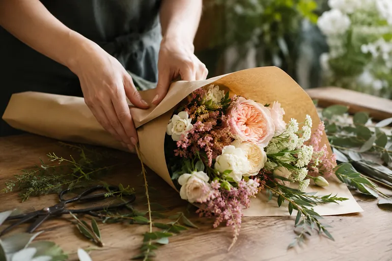 Deux mains emballent un bouquet de fleurs de saison dans du papier kraft sur un comptoir de bois, à côté de tiges coupées et d'une paire de ciseaux.