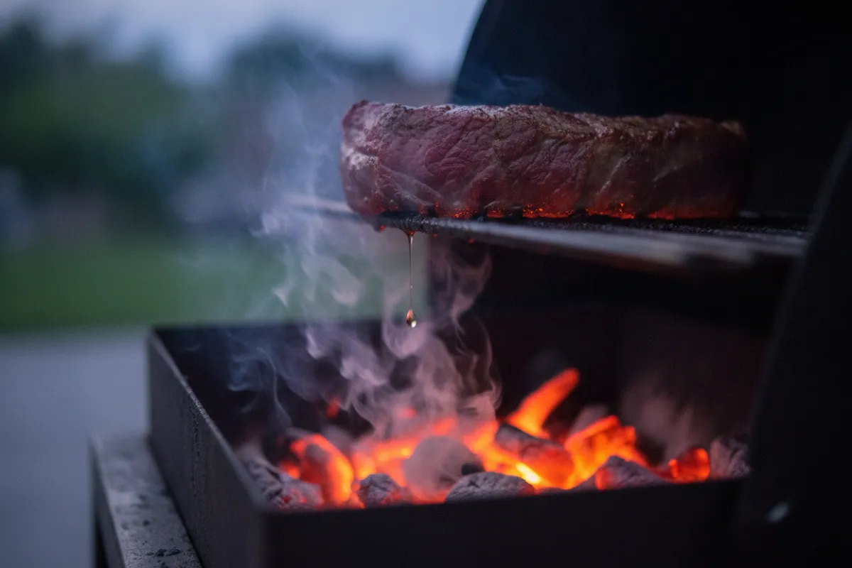 Une pièce de viande en train de cuire sur la grille d'un barbecue au-dessus de braises rougeoyantes, un filet de fumée remontant à travers la grille.